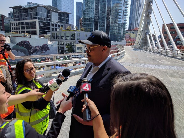 Nuru addressing the press with the bus bridge to the Bay Bridge behind him