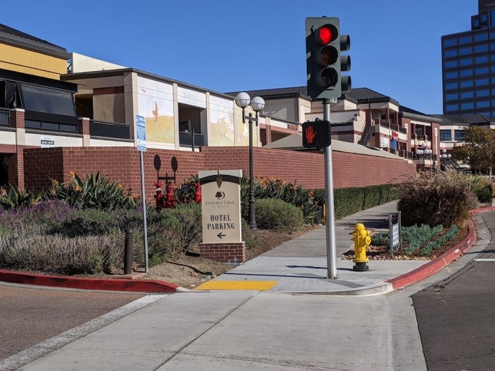 To get from the Hazard Center trolley station to this shopping center, you have to hit a beg button to get across the entrance to a parking lot, dramatic evidence of the over prioritization of cars. Photo: Streetsblog/Rudick